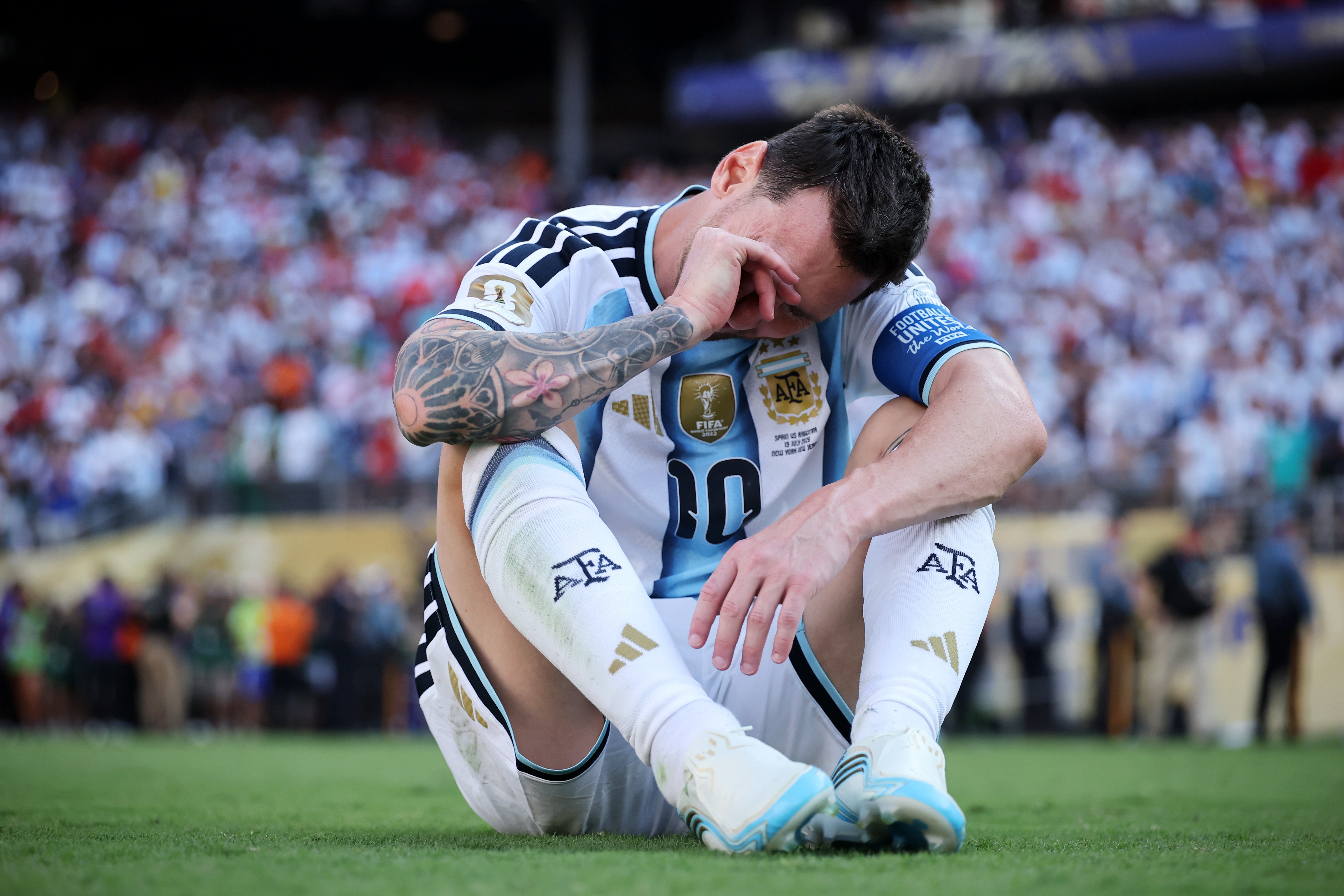 Jogador de futebol Lionel Messi, com camisa da Argentina, sentado no gramado com a cabe&ccedil;a baixa e m&atilde;o no rosto, expressando tristeza. Ao fundo, torcida emba&ccedil;ada