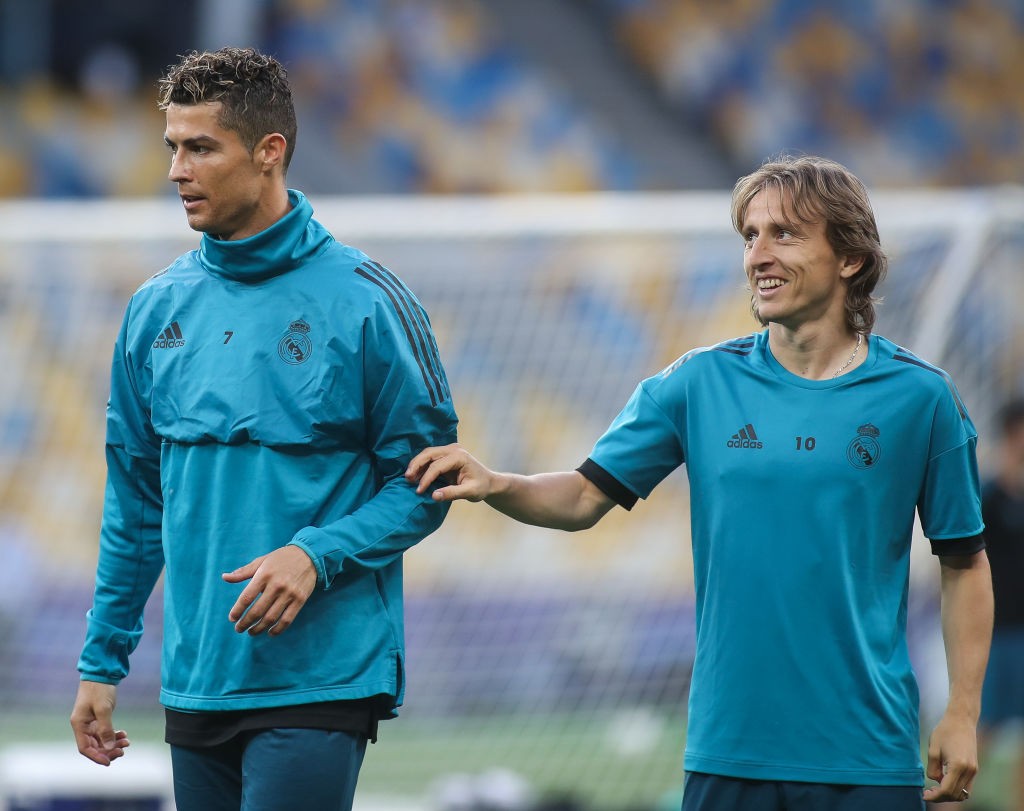 Cristiano Ronaldo, &agrave; esquerda, com agasalho azul-petr&oacute;leo de gola alta, e Luka Modric, &agrave; direita, com camisa azul-petr&oacute;leo de manga curta, sorrindo e com a m&atilde;o no ombro de Ronaldo, em um campo de futebol