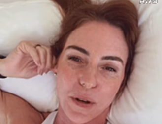 Mulher deitada em travesseiro branco, com cabelo ruivo espalhado, olhando para cima com expressão cansada. Ela tem sardas e veste uma regata branca.