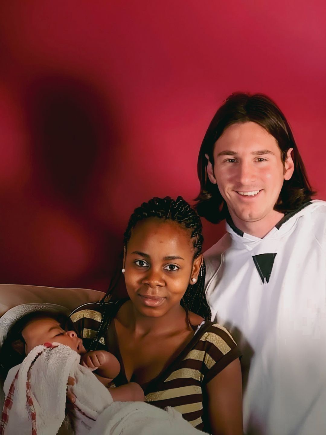 Uma mulher negra com tran&ccedil;as e blusa listrada segura um beb&ecirc; dormindo, enquanto um homem branco de cabelo comprido e camisa branca sorri ao lado, contra um fundo vermelho