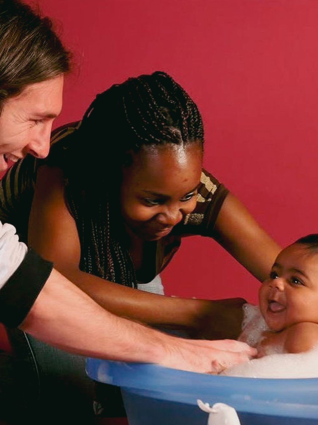Um casal sorridente, um homem branco e uma mulher negra com tran&ccedil;as, dando banho em um beb&ecirc; risonho numa banheira azul com espuma, contra um fundo vermelho
