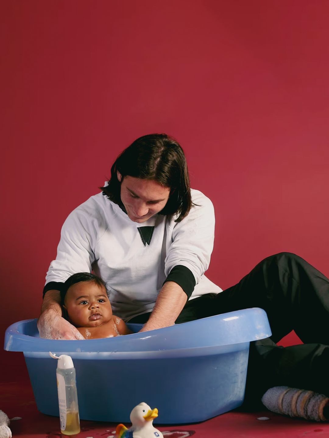 Pessoa de cabelos escuros e compridos, vestindo moletom branco e cal&ccedil;a preta, dando banho em um beb&ecirc; de pele escura em uma banheira azul. O beb&ecirc; est&aacute; com espuma no corpo e olha para frente. H&aacute; um pato de borracha e um frasco de sabonete l&iacute;quido no ch&atilde;o vermelho.