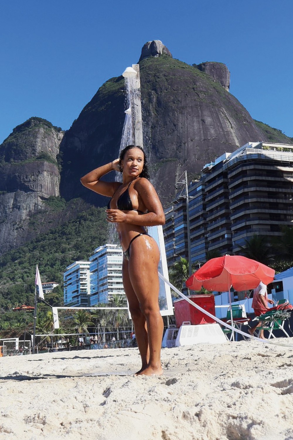 Mulher de pele morena e cabelos molhados, vestindo biqu&iacute;ni preto, toma ducha na praia. Ao fundo, montanhas rochosas, pr&eacute;dios e c&eacute;u azul
