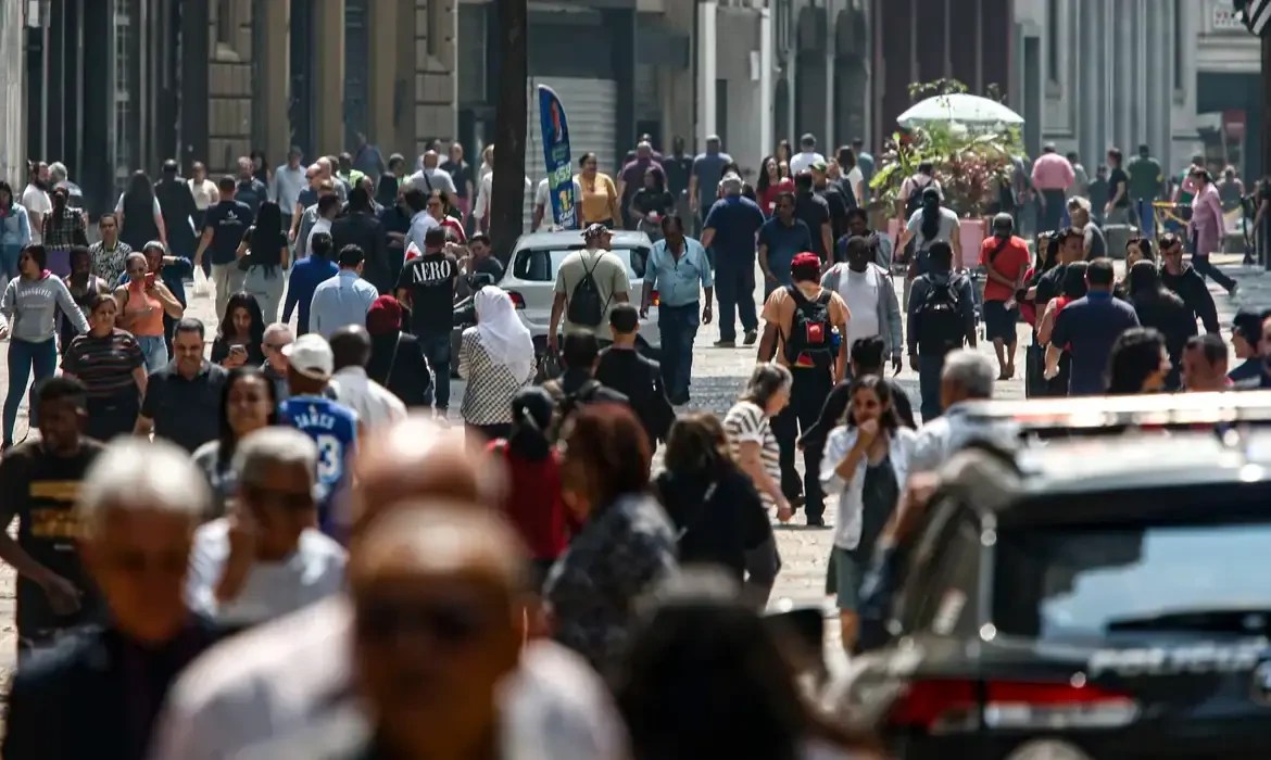 Multidão de pessoas diversas caminhando em uma rua movimentada sob luz solar intensa, com edifícios e carros ao fundo.