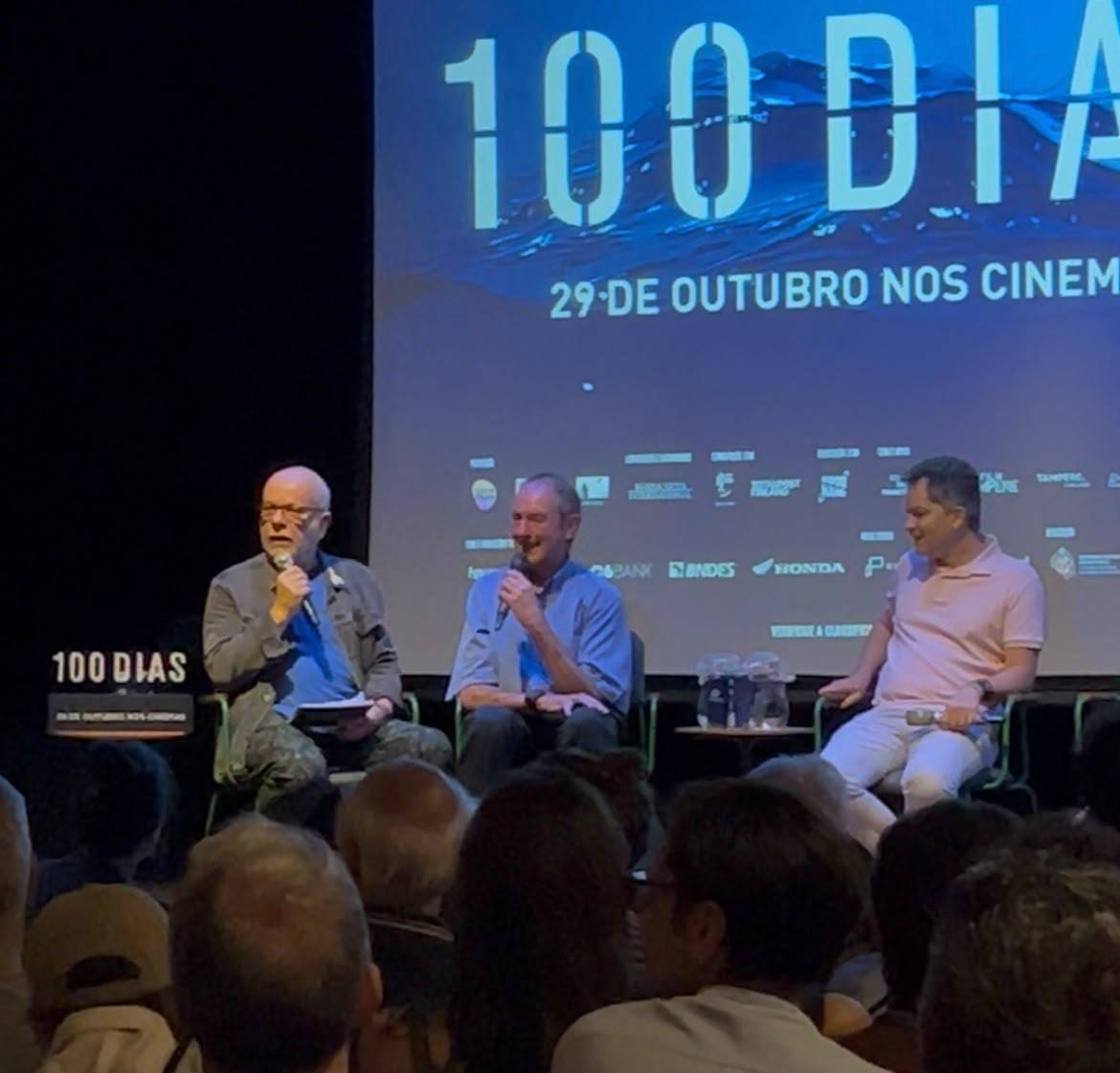 Três homens sentados em cadeiras no palco, conversando com microfones. Um telão ao fundo exibe 100 DIAS e 29 DE OUTUBRO NOS CINEMAS, com logos de patrocinadores. Uma plateia assiste de frente