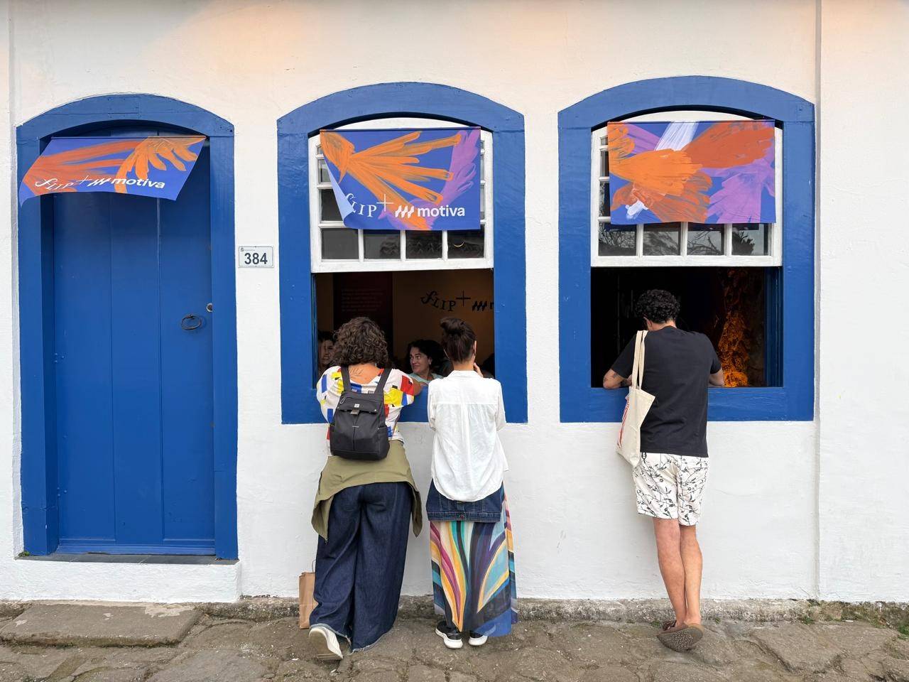 Três pessoas observam através de janelas com molduras azuis em uma fachada branca. A porta azul ao lado tem o número 384 e uma faixa SIP motiva.