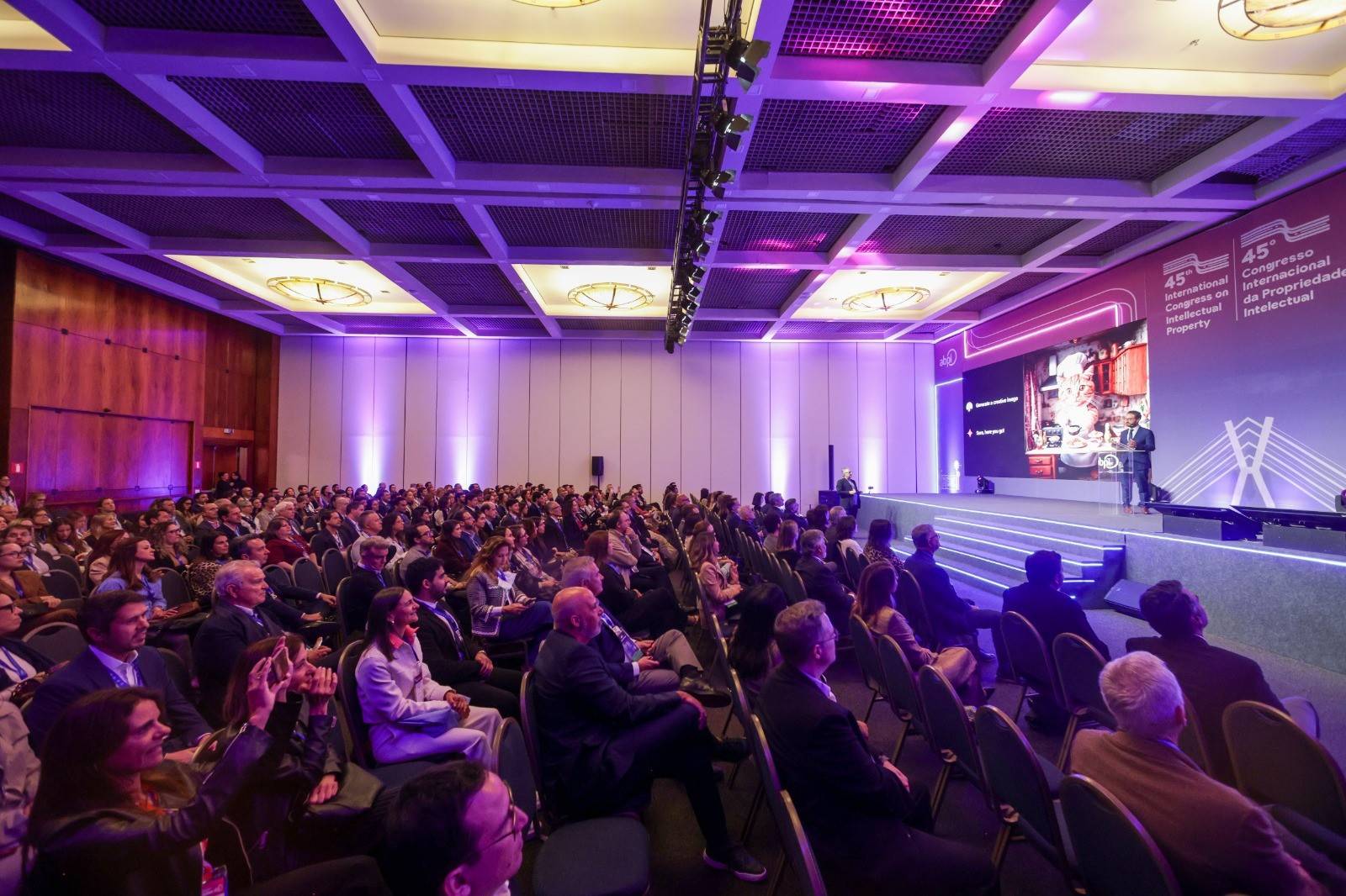 Auditório lotado com centenas de pessoas assistindo a um homem de terno azul falando em um palco iluminado por luzes roxas. Ao fundo, um telão exibe 45º Congresso Internacional de Propriedade Intelectual