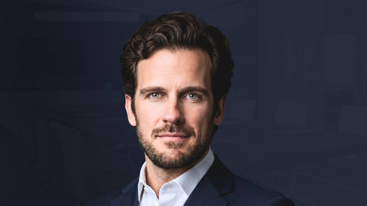 Homem branco de barba e cabelo castanho encaracolado, vestindo terno azul-marinho e camisa branca, olhando para frente com expressão séria, em fundo azul escuro com contornos de escritório desfocados