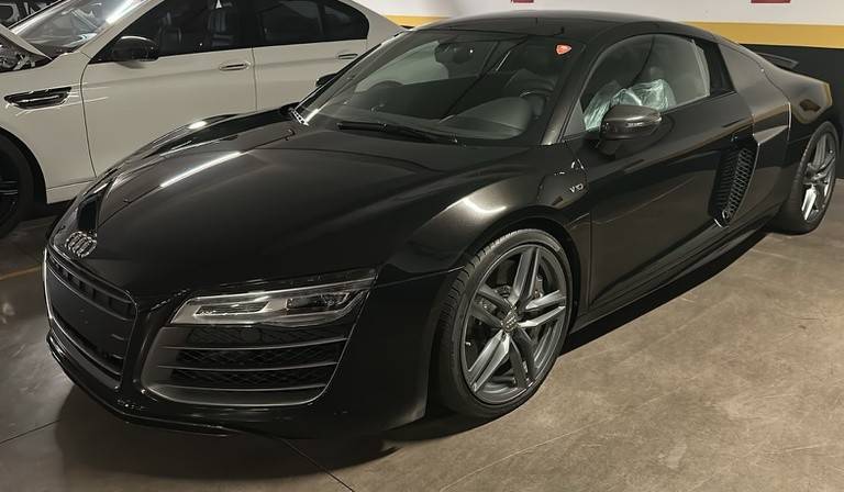 Carro esportivo Audi R8 preto brilhante estacionado em uma garagem, com um veículo branco parcialmente visível ao fundo