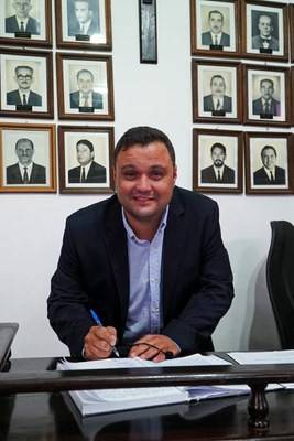 Homem de pele clara, camisa azul e blazer escuro, sorrindo enquanto assina documentos em uma mesa de madeira. Ao fundo, uma parede branca com vários retratos emoldurados de homens em preto e branco