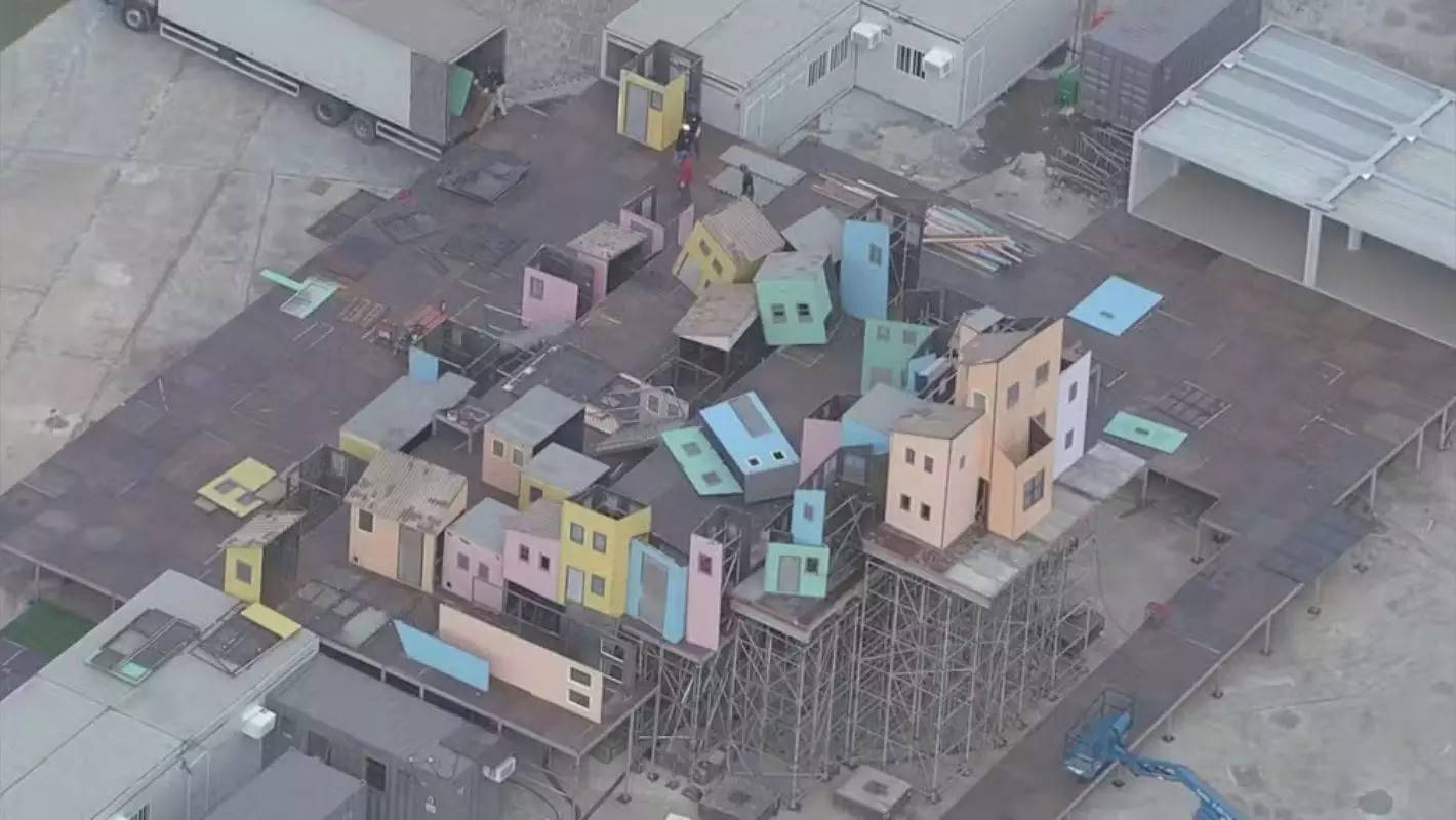 Vista aérea de um cenário de filmagem com pequenas casas coloridas, algumas inclinadas ou empilhadas, sobre plataformas e andaimes. Um caminhão branco está estacionado à esquerda, e estruturas modulares cinzas aparecem ao fundo, sugerindo um ambiente de construção ou estúdio.