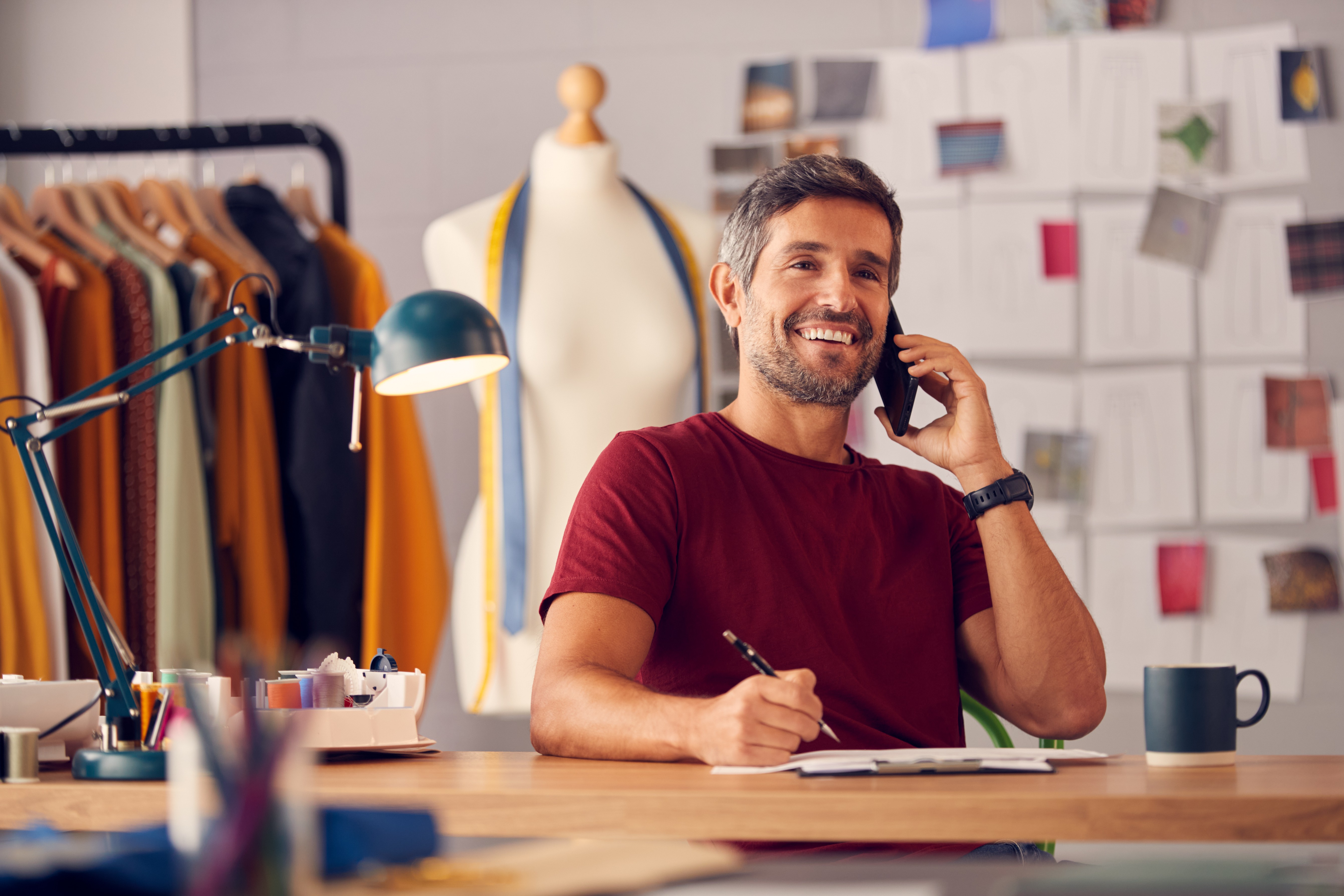 Homem barbudo de meia-idade, sorrindo, veste camiseta vinho, fala ao celular e anota em uma prancheta sobre uma mesa de madeira. Ao fundo, um manequim de costura com fita m&eacute;trica, um cabideiro com roupas e um mural de inspira&ccedil;&otilde;es. Uma lumin&aacute;ria verde ilumina a cena.