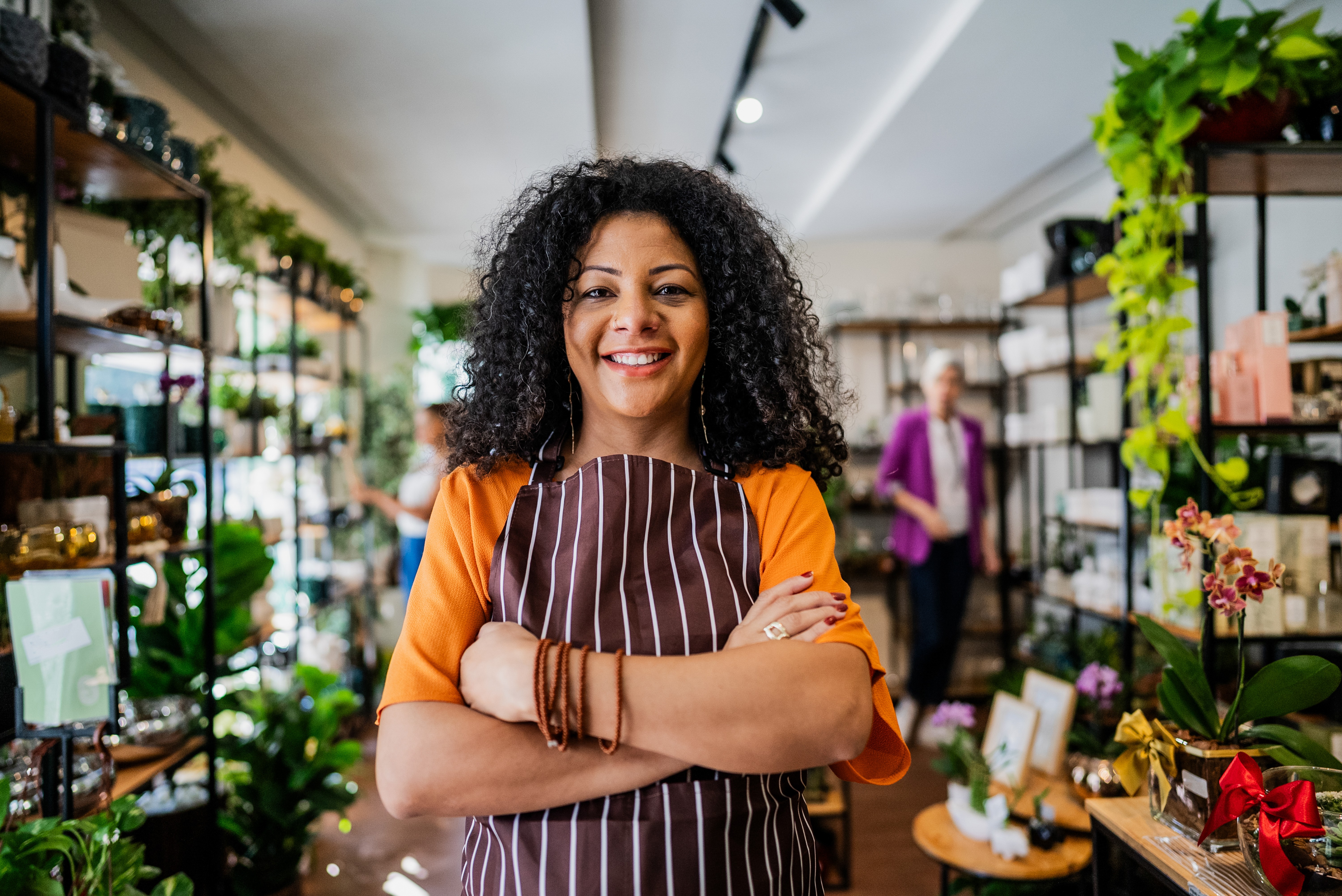 Mulher negra de cabelos cacheados, sorrindo, usando avental listrado marrom e blusa laranja, bra&ccedil;os cruzados, em uma loja com prateleiras cheias de plantas e produtos. Ao fundo, pessoas desfocadas.