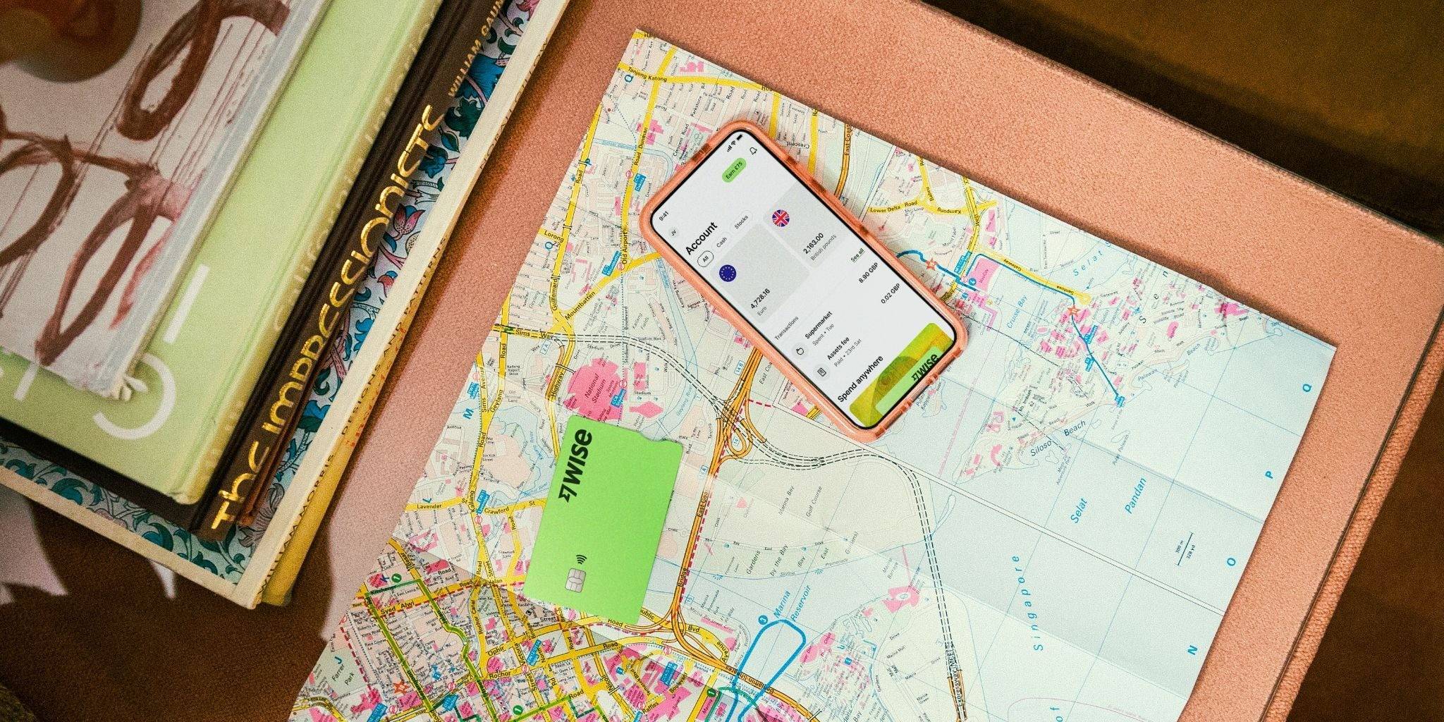 Um smartphone com capa rosa exibe a tela de um aplicativo financeiro sobre um mapa de ruas. Ao lado, um cartão verde da Wise e uma pilha de livros.