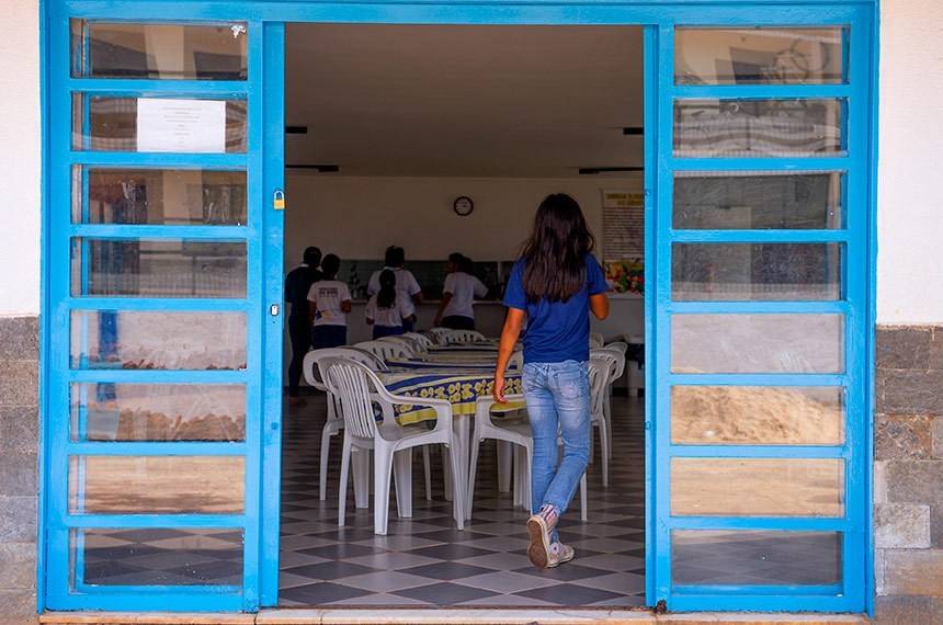Uma jovem de cabelo escuro e comprido, vestindo camiseta azul e calça jeans, entra em um refeitório simples com piso xadrez. Ela caminha em direção a mesas com cadeiras brancas de plástico, onde outras pessoas estão de pé ao fundo