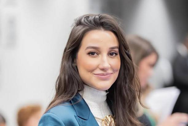 Mulher jovem de cabelos castanhos longos e ondulados, sorrindo levemente, vestindo um blazer azul-petróleo sobre uma blusa de gola alta branca e um colar dourado. O fundo está desfocado, com outras pessoas indistintas.