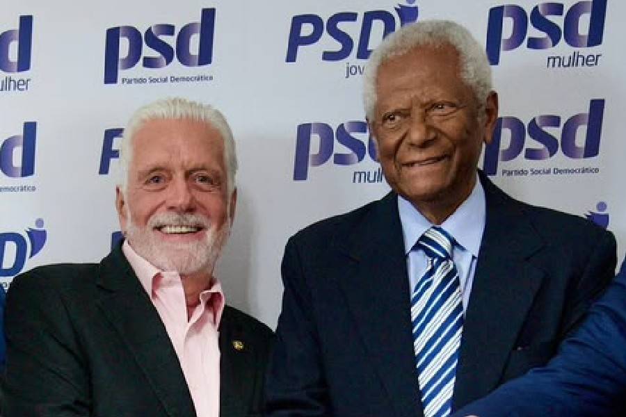 Dois homens sorrindo, um branco com barba grisalha e camisa rosa, e outro negro de terno azul e gravata listrada, em frente a um painel com o logo do PSD
