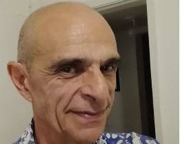 Homem calvo de pele clara, com rugas na testa e ao redor dos olhos, sorri levemente. Veste camisa estampada em tons de azul e branco, com fundo claro.