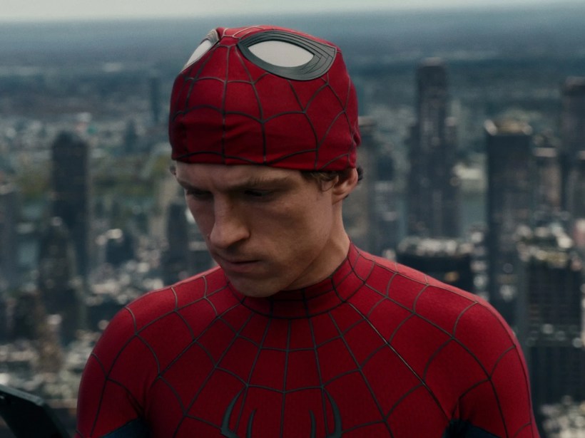 Homem-Aranha, com parte da máscara levantada revelando seu rosto sério, olha para baixo enquanto segura um celular. Ele veste o traje vermelho e azul, com a cidade ao fundo