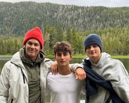 Tom Brady, sorrindo, com gorro vermelho e jaqueta cinza, abraça dois jovens, um com camiseta branca e outro com gorro azul e moletom cinza, em frente a um lago e floresta densa