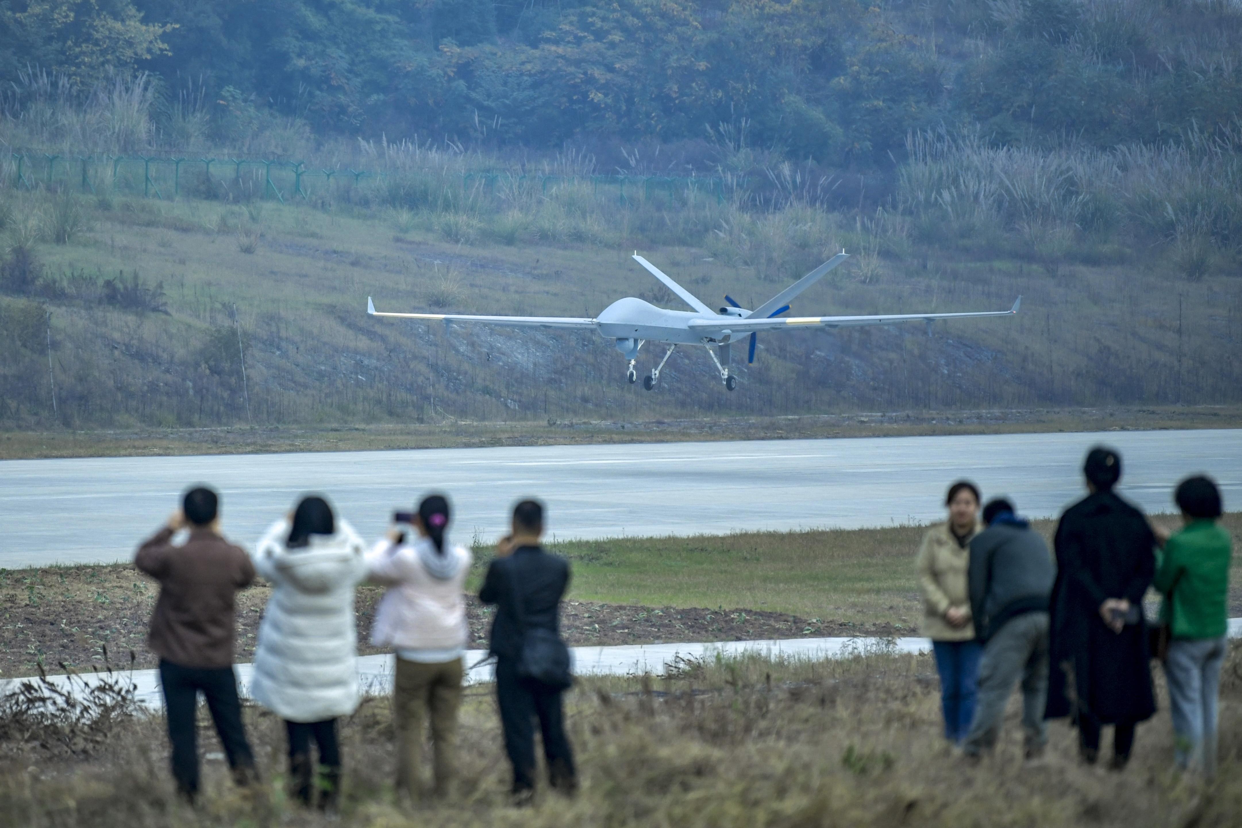 Um drone militar branco com asas longas e trem de pouso estendido se aproxima de uma pista de pouso, visto por um grupo de pessoas em primeiro plano, algumas fotografando. O cen&aacute;rio &eacute; um campo com vegeta&ccedil;&atilde;o rasteira e colinas ao fundo