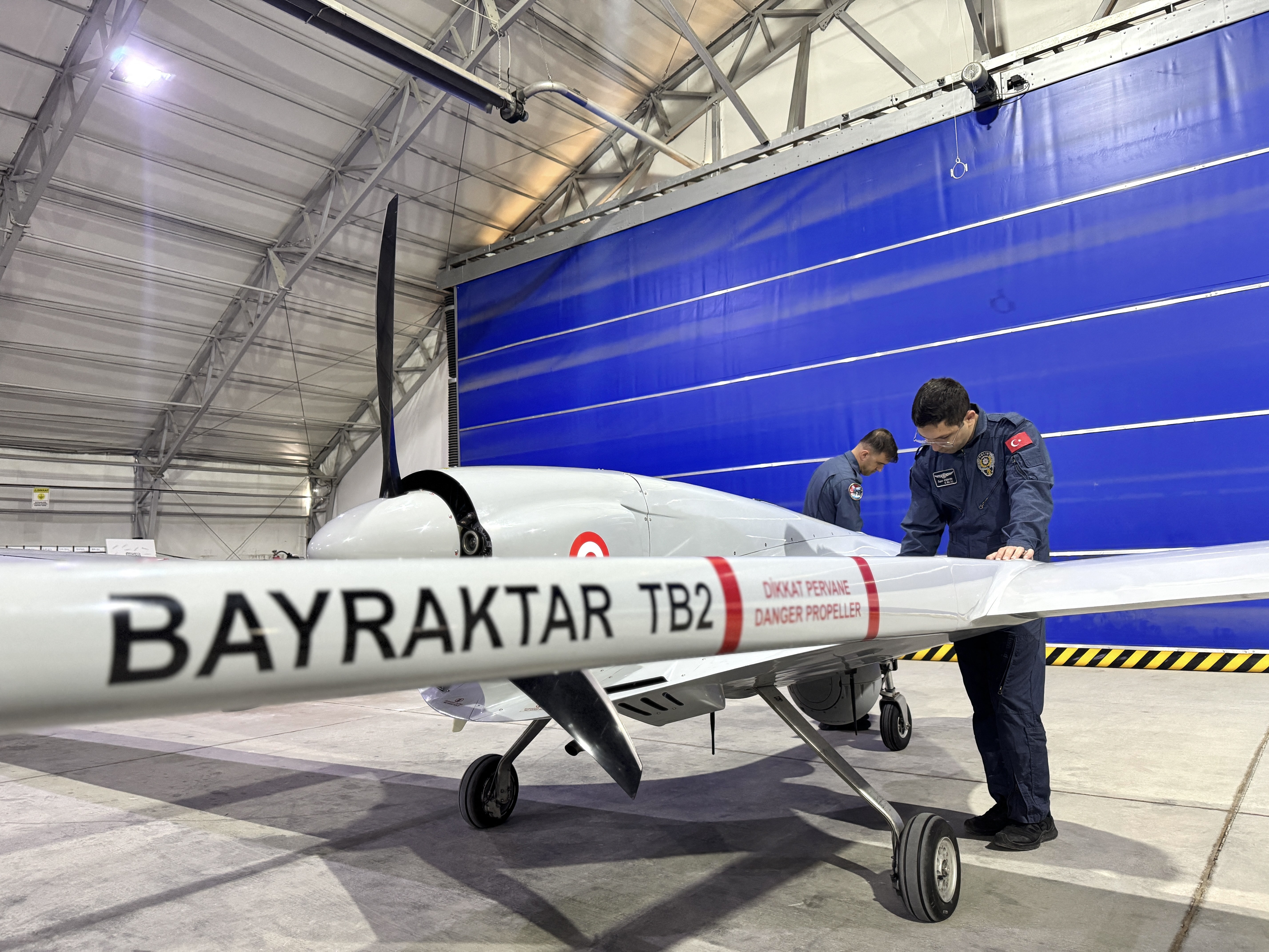 Dois homens de uniforme azul inspecionam um drone militar branco Bayraktar TB2 com h&eacute;lice traseira, dentro de um hangar com teto met&aacute;lico e parede azul