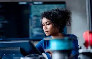 Mulher jovem com cabelo cacheado escuro, vestindo blusa azul, concentrada enquanto olha para uma tela ou tablet em um ambiente de trabalho com monitores e equipamentos eletr&ocirc;ncos ao fundo
