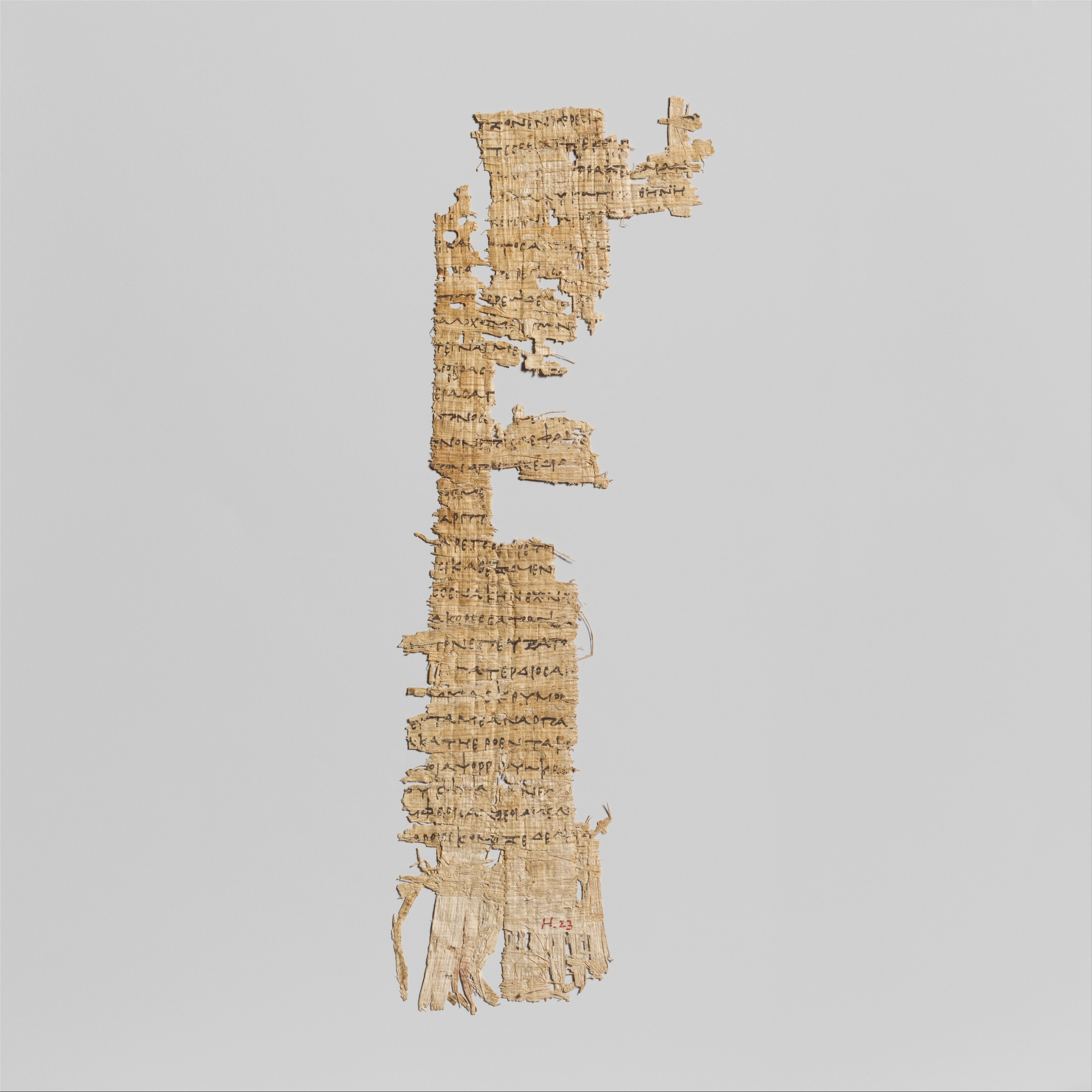 Fragmento vertical de papiro antigo, cor bege claro, com texto em escrita escura e uma cruz no topo. As bordas s&atilde;o irregulares e desfiadas, revelando a textura fibrosa do material. O fundo &eacute; cinza claro e liso