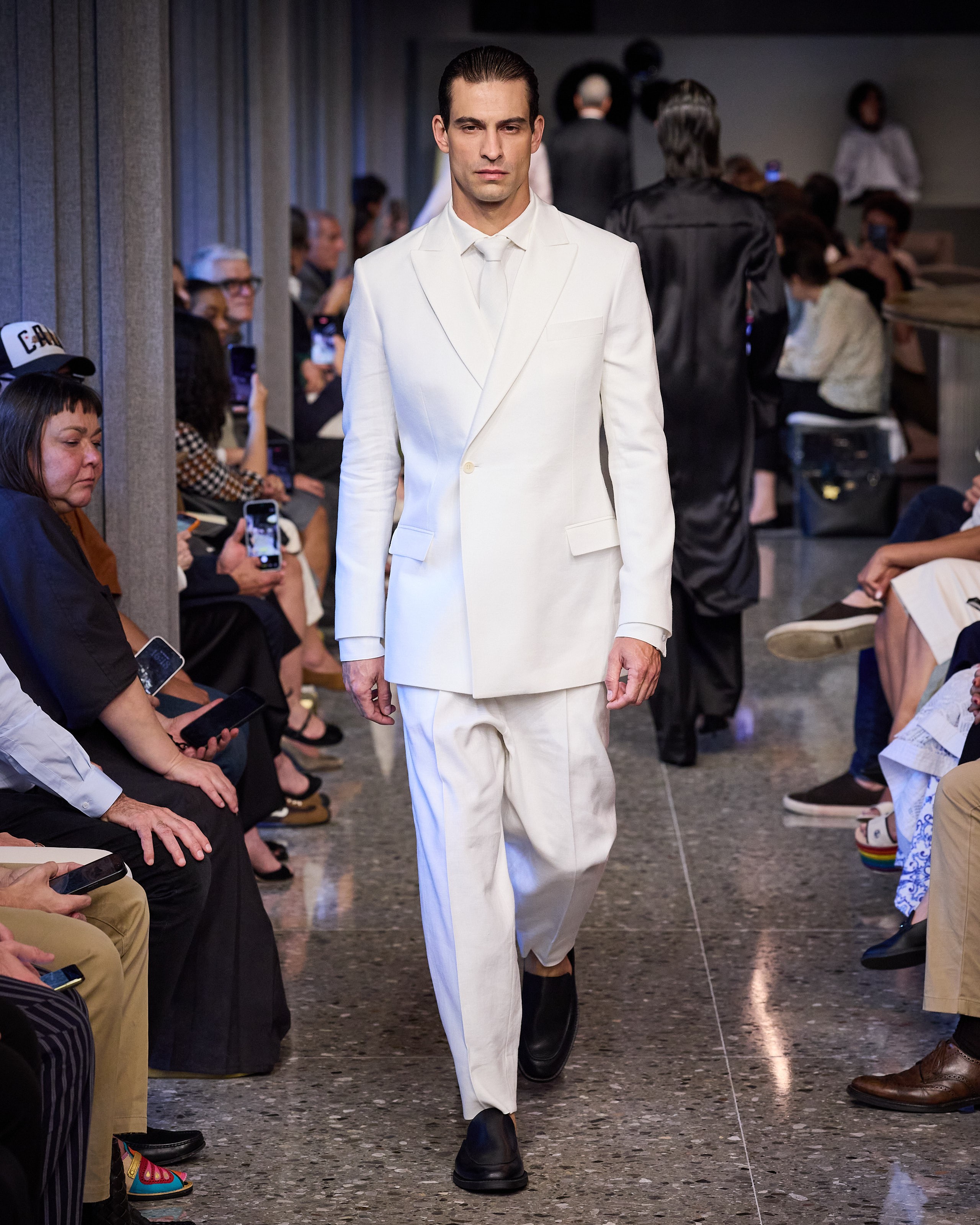 Modelo masculino com cabelo penteado para tr&aacute;s, vestindo um terno branco monocrom&aacute;tico com gravata, desfilando em uma passarela. Pessoas sentadas assistem ao desfile, algumas segurando celulares