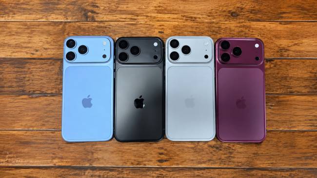 Quatro smartphones de costas, alinhados horizontalmente sobre uma superf&iacute;cie de madeira. Da esquerda para a direita, os celulares s&atilde;o azul-claro, preto, cinza-claro e roxo-escuro. Todos possuem o logo da Apple no centro e tr&ecirc;s lentes de c&acirc;mera na parte superior esquerda