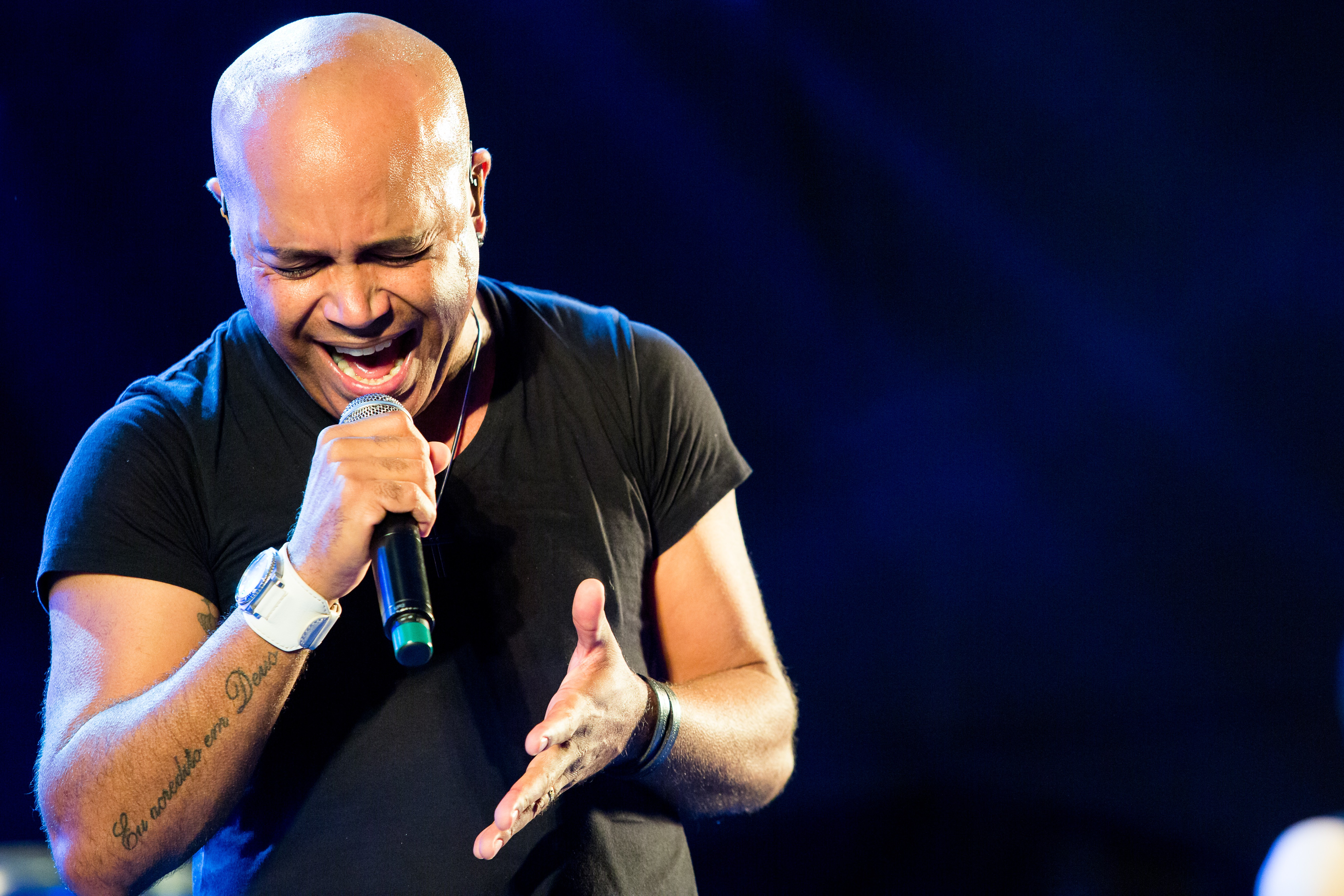 Cantor careca de pele morena, vestindo camiseta preta, cantando com microfone na mão esquerda e braço direito estendido, em palco escuro com iluminação azul. Ele tem uma tatuagem no antebraço esquerdo e usa um relógio branco. Sua boca está aberta e os olhos fechados, transmitindo emoção