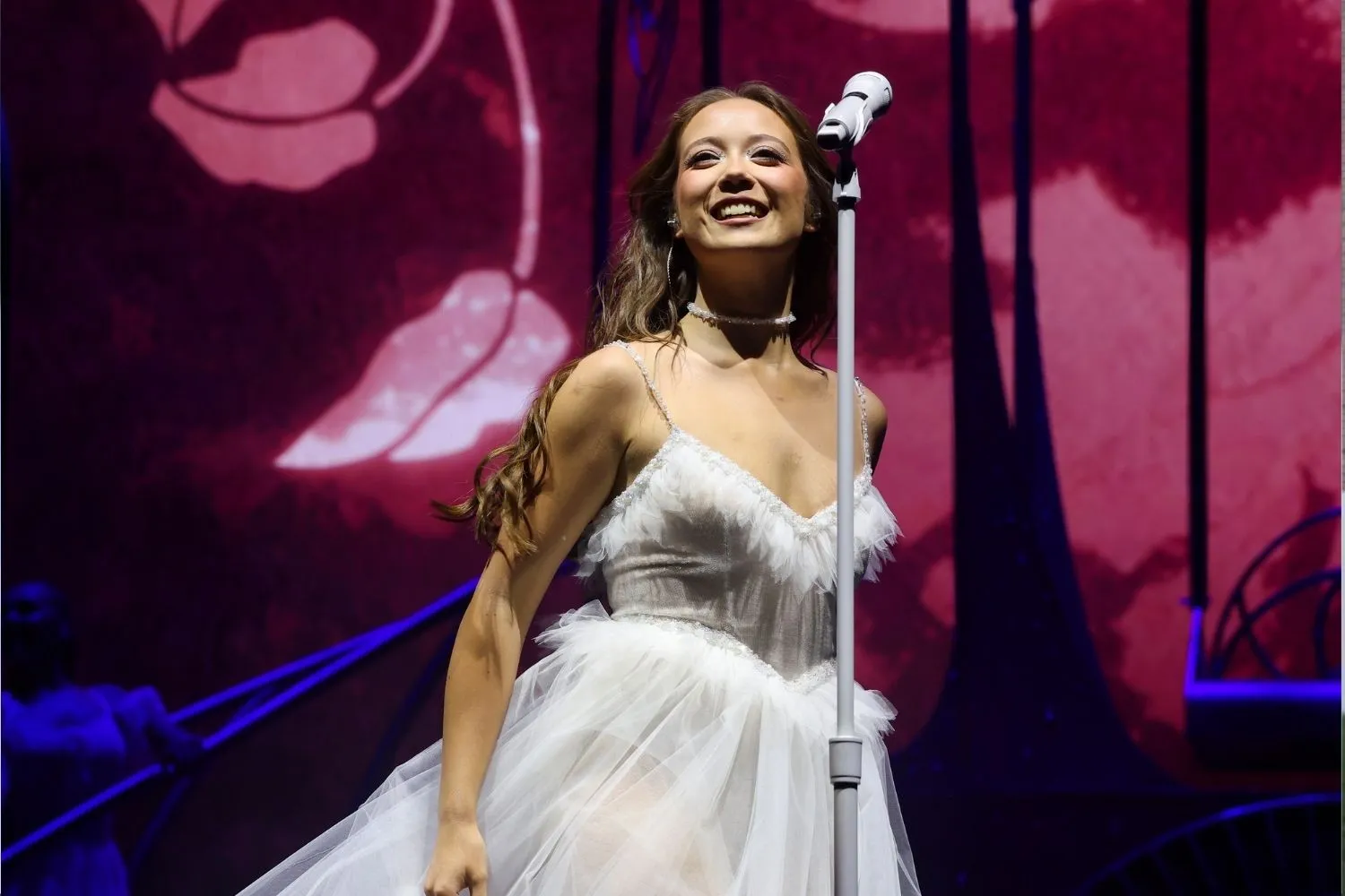 Cantora sorridente de cabelos castanhos e vestido branco de tule, com alças finas e decote em V, em palco escuro com projeções de flores rosadas e um microfone à sua frente