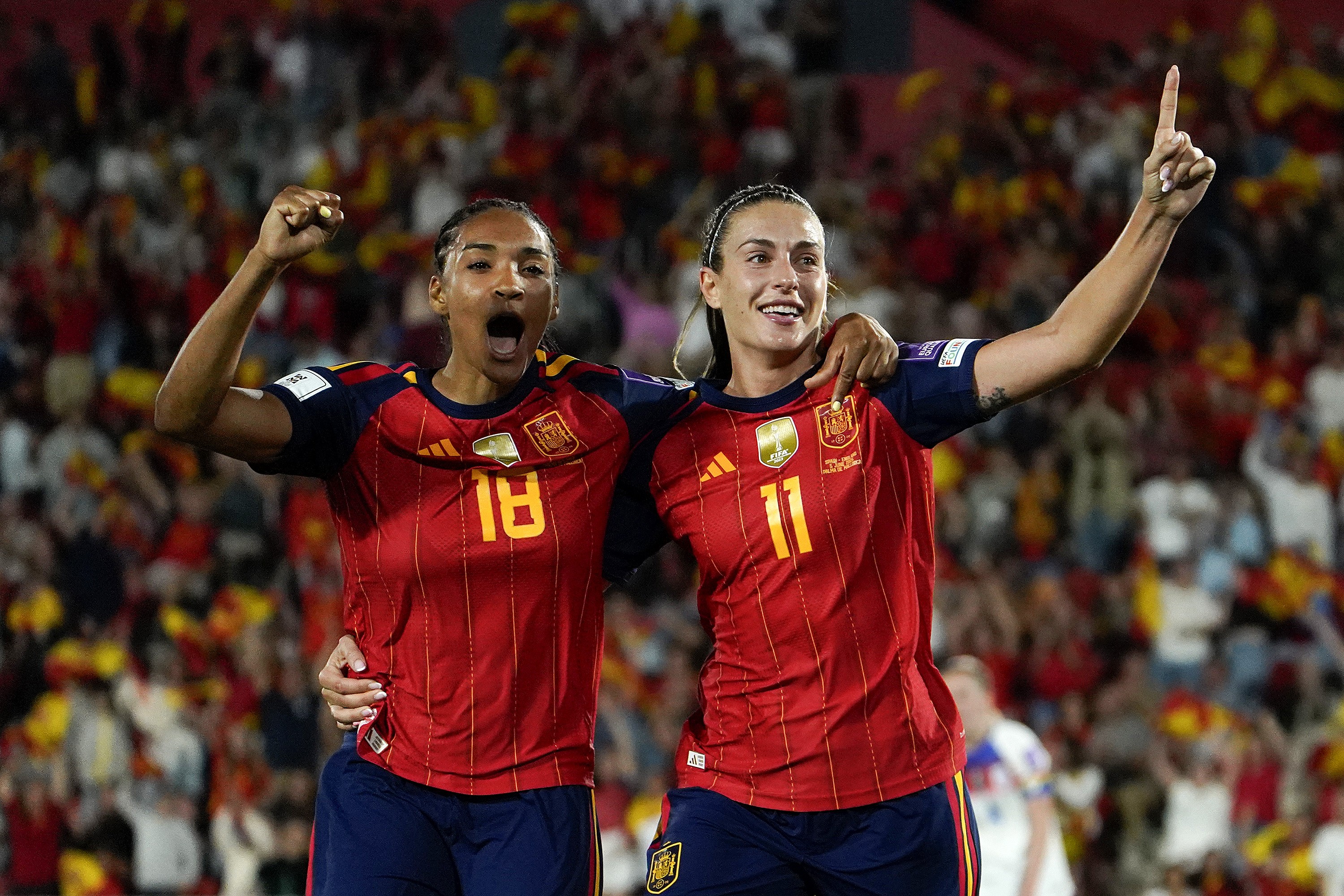 Duas jogadoras de futebol espanholas, com camisas vermelhas e detalhes azuis, celebram em campo. A da esquerda, com camisa 18, tem o punho cerrado e boca aberta em grito de alegria. A da direita, com camisa 11, sorri e aponta o dedo indicador para cima, com o braço da colega sobre seu ombro. Torcida borrada ao fundo.
