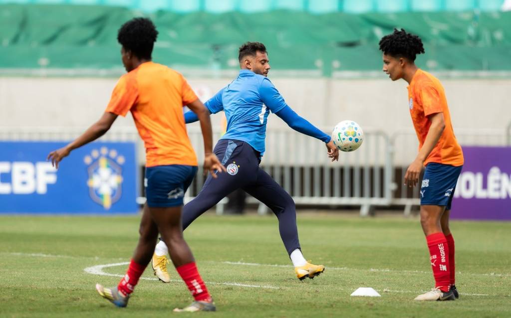 Tr&ecirc;s jogadores de futebol em campo verde. Um homem de camisa azul e cal&ccedil;a escura domina a bola com a m&atilde;o, enquanto dois jogadores de camisa laranja e shorts azuis observam. Um deles est&aacute; de costas, e o outro, &agrave; direita, olha para a bola. Ao fundo, faixas com o logo da CBF e a palavra VIOL&Ecirc;NCIA