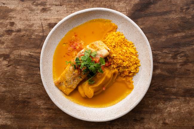 Prato de peixe com molho amarelo, pur&ecirc; de ab&oacute;bora e farofa crocante, decorado com cheiro-verde e pimenta picada, sobre uma mesa de madeira r&uacute;stica
