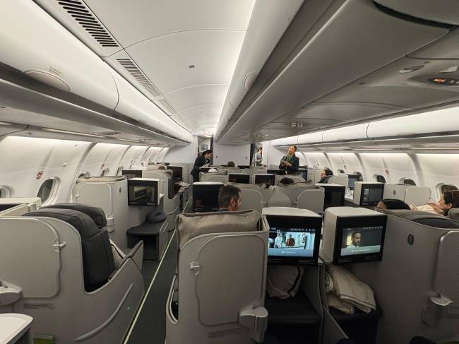 Interior de um avi&atilde;o com assentos de classe executiva, onde passageiros assistem a telas individuais. Comiss&aacute;rios de bordo circulam pelo corredor, e a ilumina&ccedil;&atilde;o suave destaca o ambiente espa&ccedil;oso e confort&aacute;vel