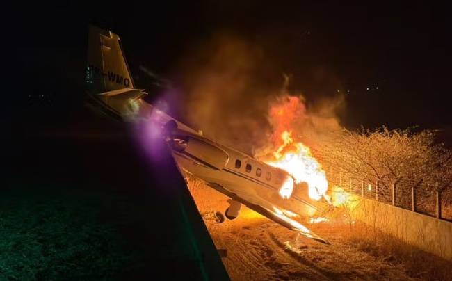 Um avi&atilde;o de pequeno porte em chamas, com fogo intenso e fuma&ccedil;a laranja, inclinado em um terreno escuro &agrave; noite. A aeronave branca tem a cauda amarela com a inscri&ccedil;&atilde;o PR-WMO e est&aacute; ao lado de uma cerca e &aacute;rvores secas, iluminadas pelo fogo. Uma luz roxa brilha no lado esquerdo da imagem, contrastando com o cen&aacute;rio escuro.