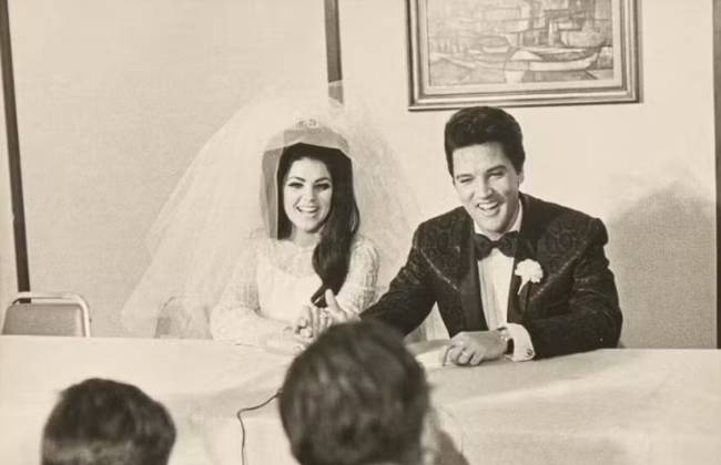 Priscilla Presley, com v&eacute;u e vestido de noiva branco, e Elvis Presley, de terno preto com brocado e gravata borboleta, sorriem sentados &agrave; mesa em seu casamento. Ao fundo, um quadro com barcos. Em primeiro plano, duas pessoas de costas observam o casal