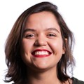 Mulher jovem, de pele clara e cabelos castanhos curtos, sorri amplamente mostrando os dentes, com batom vermelho vibrante. Ela usa uma blusa escura e está em um fundo branco.