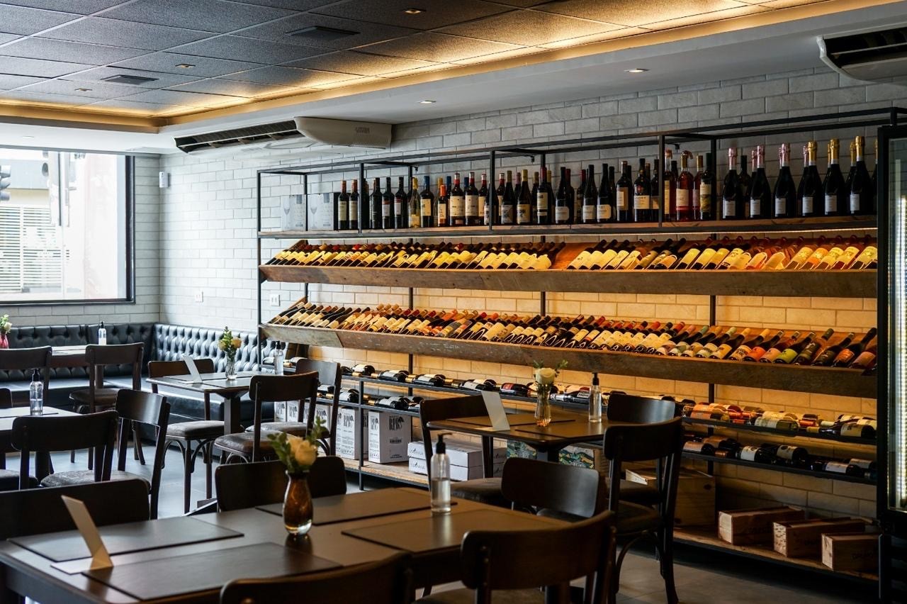 Interior de um restaurante ou adega com ilumina&ccedil;&atilde;o quente, mostrando prateleiras de madeira e metal repletas de garrafas de vinho. Mesas e cadeiras escuras ocupam o primeiro plano, com um banco estofado ao fundo, pr&oacute;ximo a uma grande janela.