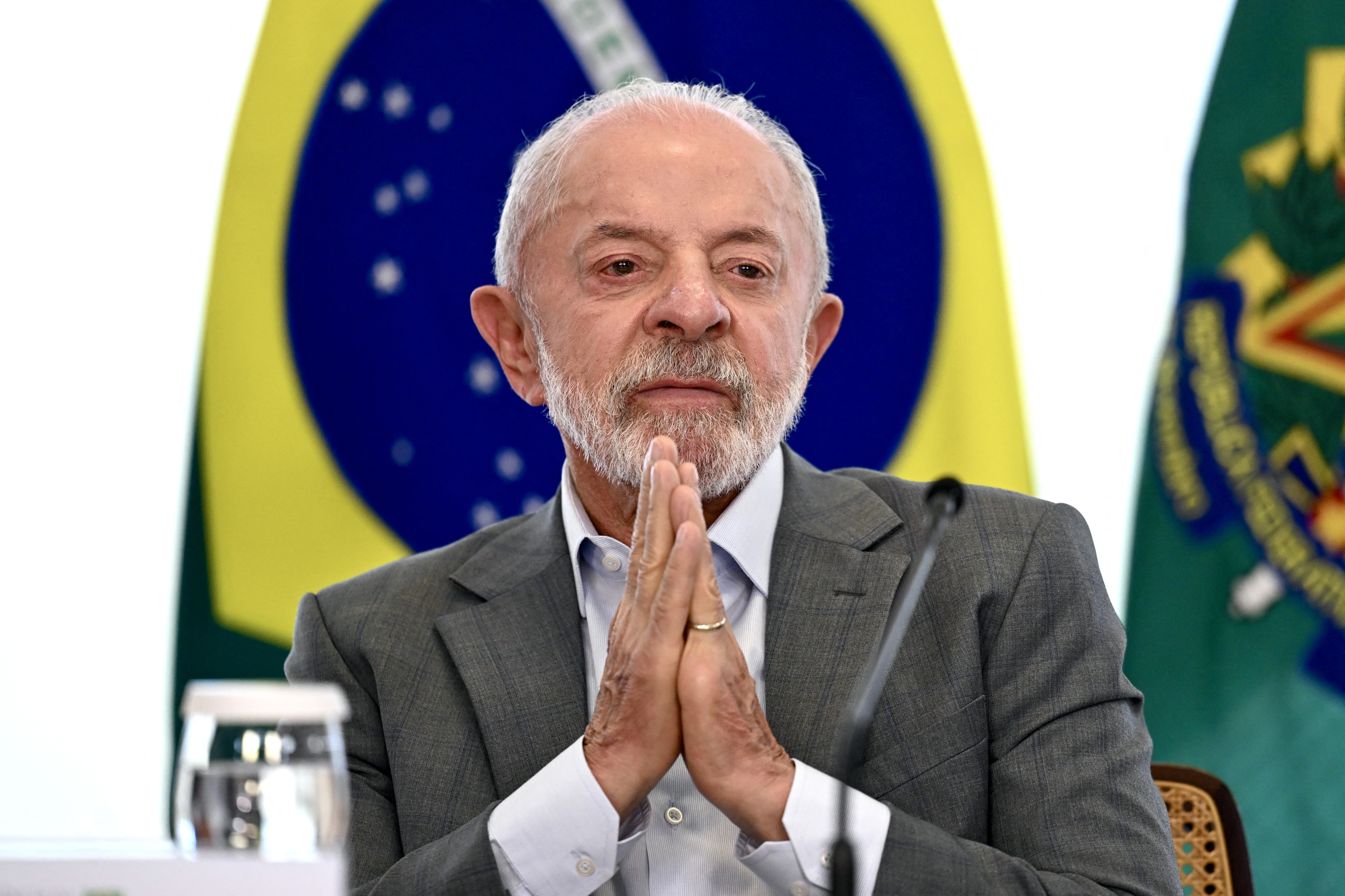 Lula, presidente do Brasil, com barba e cabelos brancos, veste terno cinza e camisa clara, mãos unidas em frente ao corpo, expressão séria. Ao fundo, bandeiras do Brasil e do Mercosul.