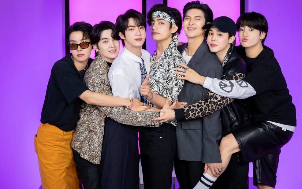 Sete homens jovens, membros do grupo BTS, posam abraçados e sorridentes contra um fundo roxo. Eles vestem roupas casuais e estilosas, com um deles usando óculos escuros e outro uma bandana na cabeça.