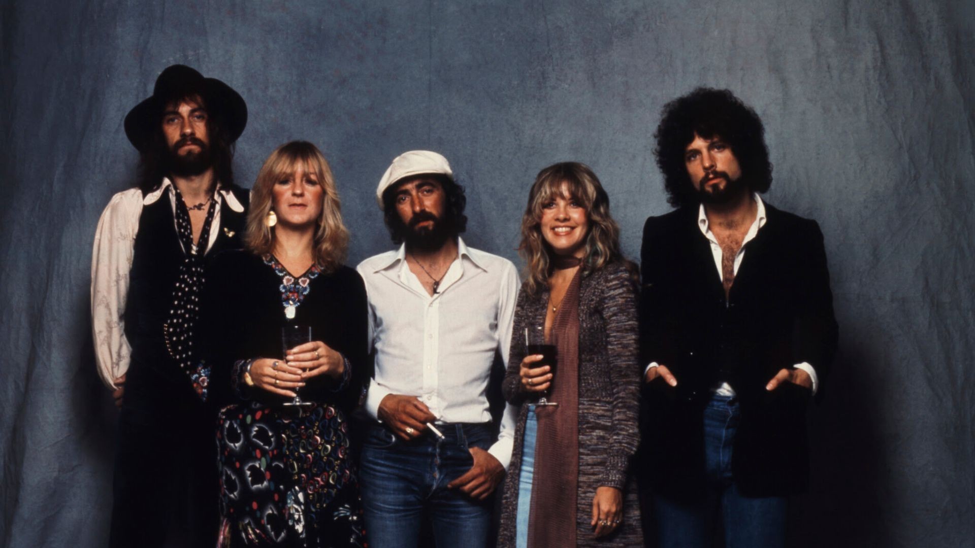 Cinco membros da banda Fleetwood Mac posam para foto, com dois homens e uma mulher à esquerda, e uma mulher e um homem à direita, todos com roupas casuais dos anos 70, em fundo cinza.