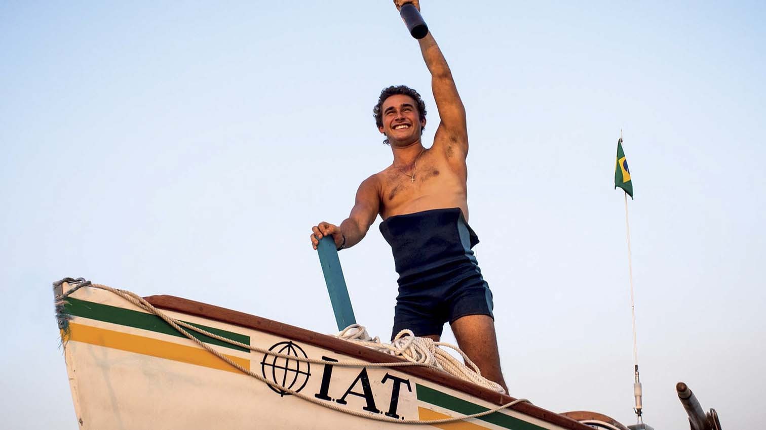 Homem sorridente, sem camisa, em pé na proa de um barco de madeira, com um remo na mão esquerda e o braço direito erguido segurando um objeto escuro. O barco tem faixas verde e amarela, e a sigla IAT com um globo. Ao fundo, céu claro e uma bandeira do Brasil tremulando.