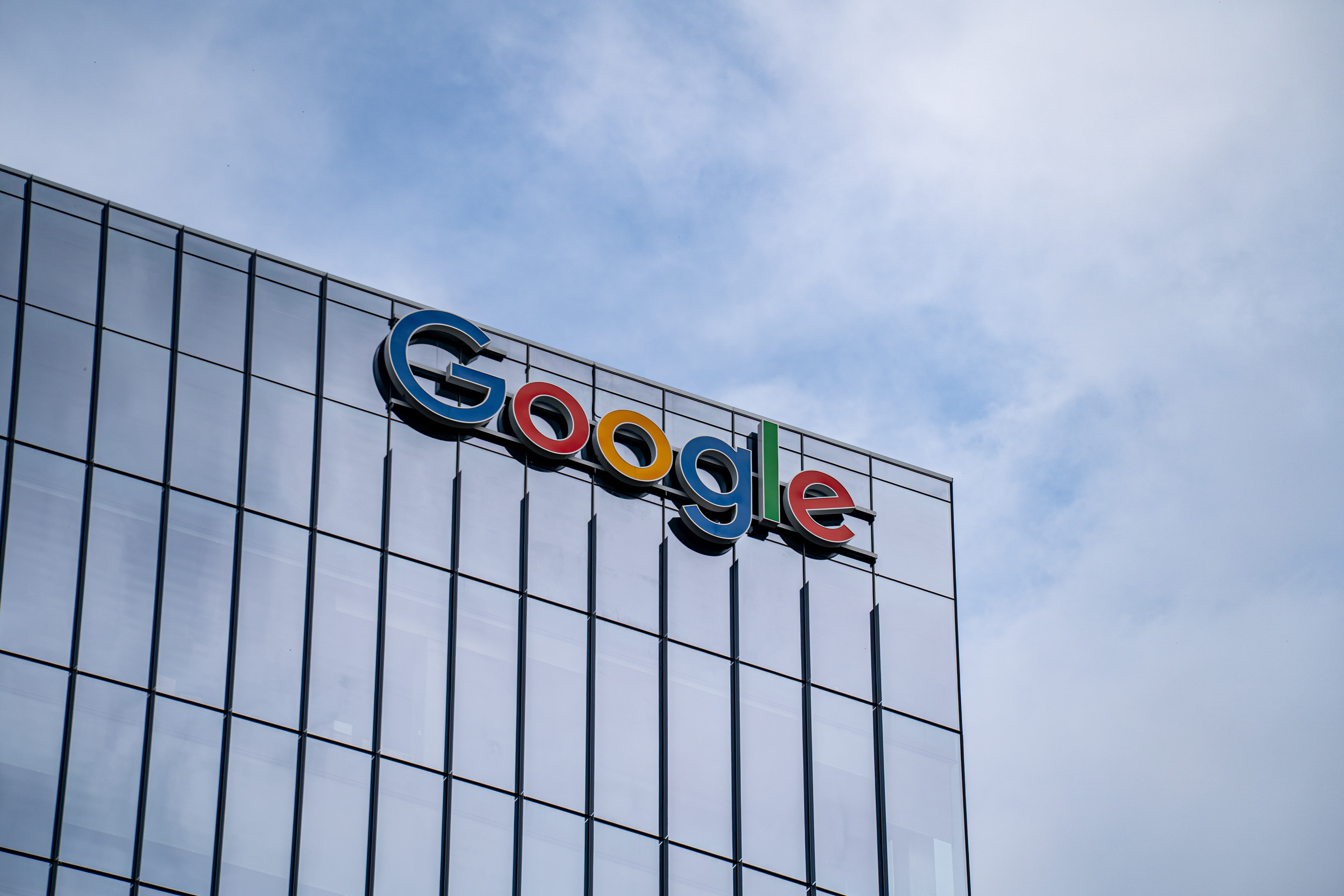 Letreiro colorido do Google no topo de um edifício moderno com fachada de vidro espelhado, sob um céu azul com nuvens esparsas