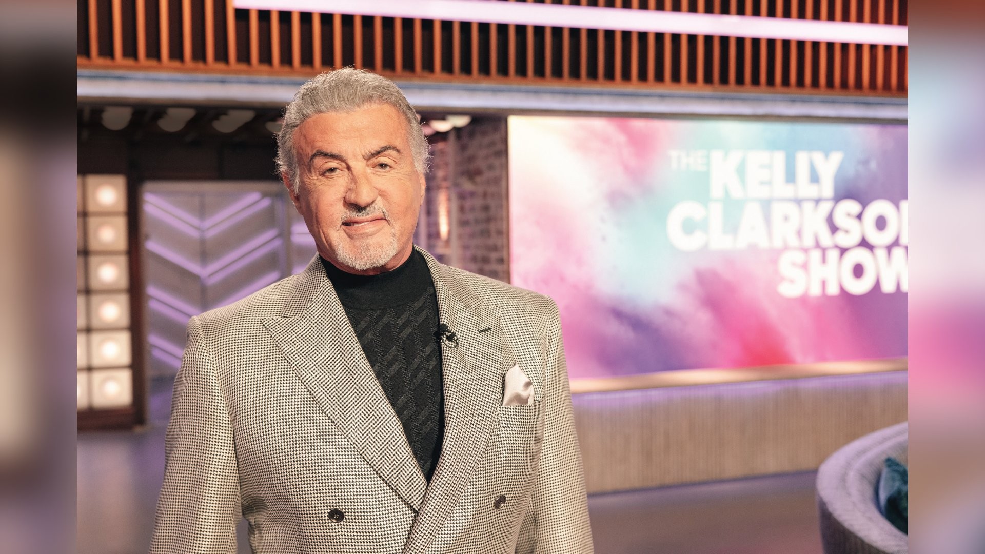 Sylvester Stallone, com cabelos grisalhos e barba, sorri levemente, vestindo um blazer xadrez cinza sobre uma blusa preta de gola alta. Ele está em um estúdio de TV, com um microfone de lapela, e ao fundo, um painel colorido exibe The Kelly Clarkson Show