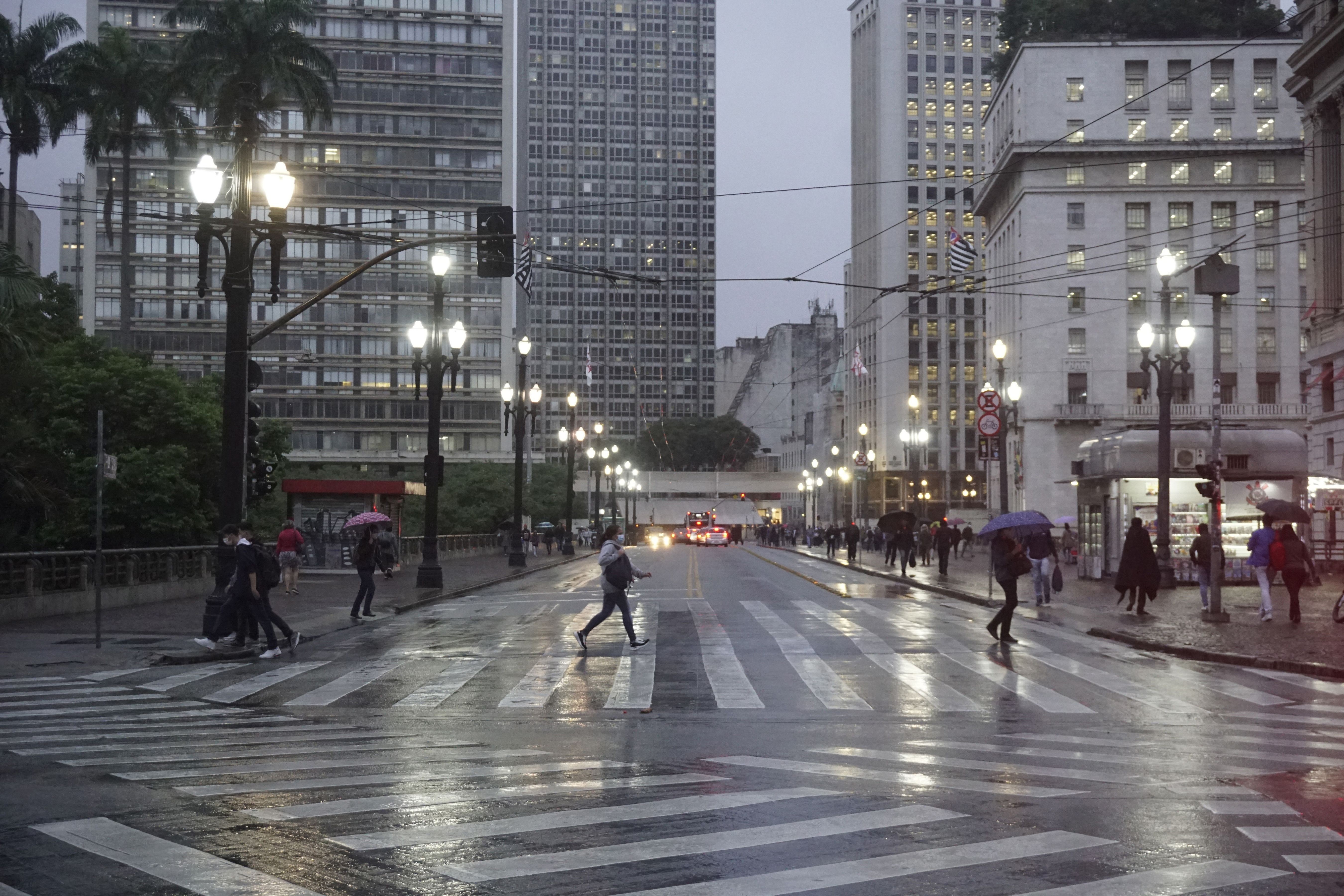 Vista noturna de uma rua molhada em cidade grande, com pedestres atravessando a faixa de segurança. Postes de luz acesos iluminam a cena, e edifícios altos com janelas iluminadas se erguem ao fundo.
