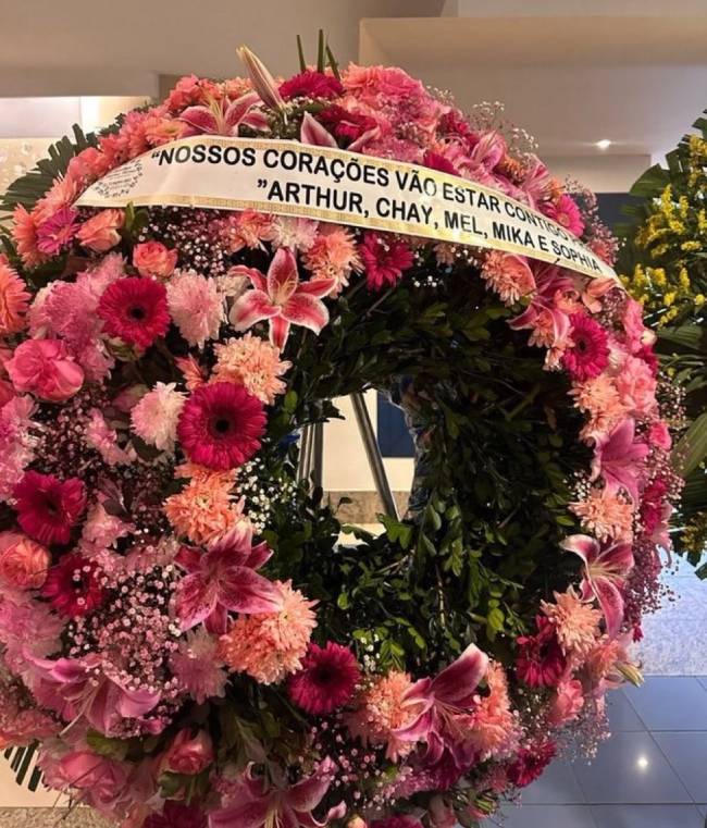 Coroa de flores f&uacute;nebre em tons de rosa, vermelho e p&ecirc;ssego, com folhagens verdes. Uma faixa branca com a frase NOSSOS CORA&Ccedil;&Otilde;ES V&Atilde;O ESTAR CONTIGO e os nomes ARTHUR, CHAY, MEL, MIKA E SOPHIA atravessa o centro